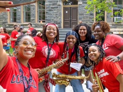 A group of women in red t-shirts, some holding brass instruments, take a selfie on the Arts Quad during Reunion 2026