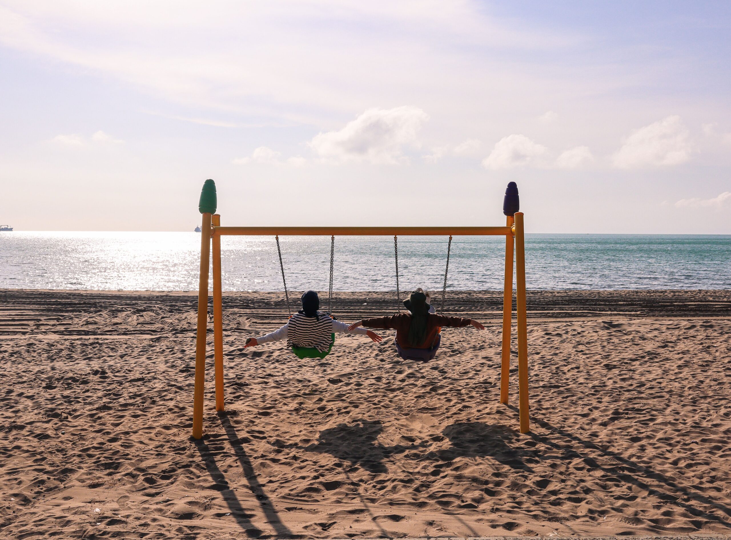 Two children on swings enjoying a sunny day at the beach with a scenic ocean view.